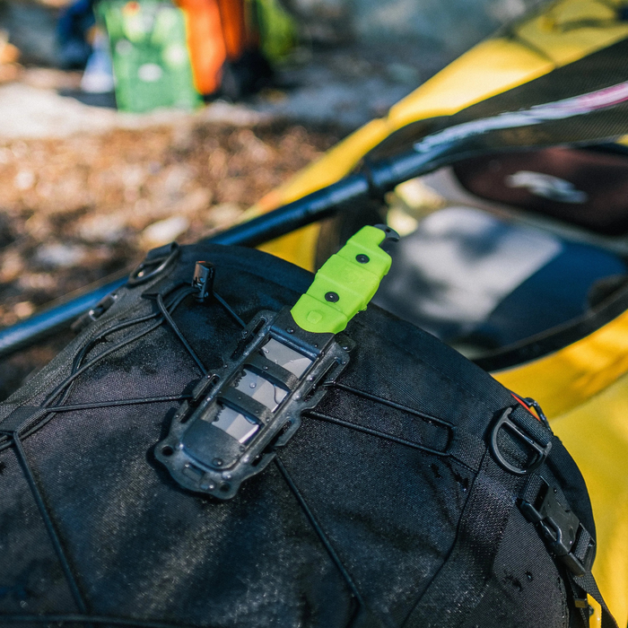 Green Gear Aid Akua River Knife Attached to the outside of a black Kayaking Gear Bag with a yellow kayak and paddle in the background 