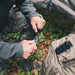 Model using a coyote coloured Gear Aid Kotu knife cutting a cord outside beside a gear bag with the quick release sheath attached